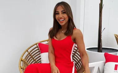A person in a red dress sitting on a wicker chair with red and white cushions, smiling.
