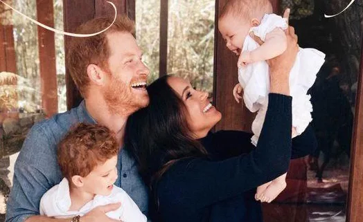 Family smiling and holding two young children, one in each parent's arms, standing in front of a window.