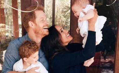 Family smiling and holding two young children, one in each parent's arms, standing in front of a window.
