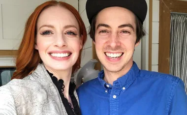 A couple smiling widely in a bright room, the woman with red hair and the man wearing a blue shirt and cap.