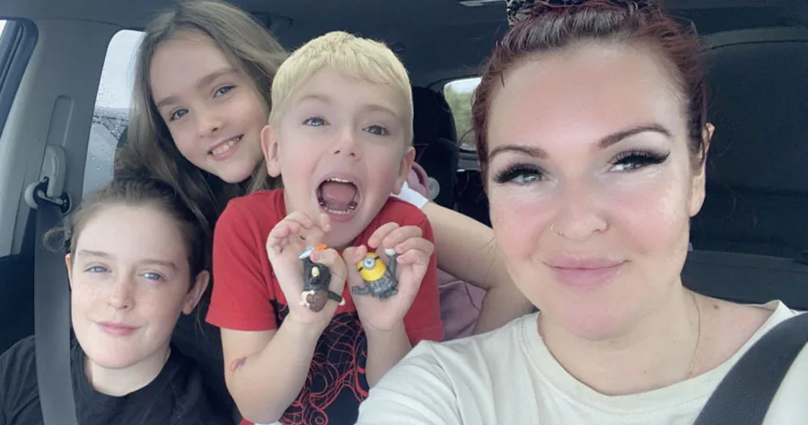 Family selfie in a car: woman in front, three kids in back, one with Minion toys.