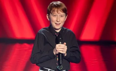 Young boy with a microphone on stage, wearing a black shirt, in front of red lights.