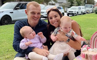 A couple smiles while holding two babies outdoors on a grassy area, near a parked car. A pink cake is in the foreground.