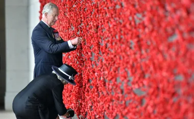 Two individuals place poppies at a memorial wall covered in red flowers during an ANZAC Day tribute.
