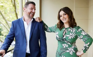 Two people smiling, one in a blue suit and the other in a green floral dress, standing on a balcony with trees behind.