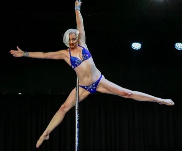 A person in a blue outfit performs a pole dance, demonstrating strength and balance with one arm and leg extended.