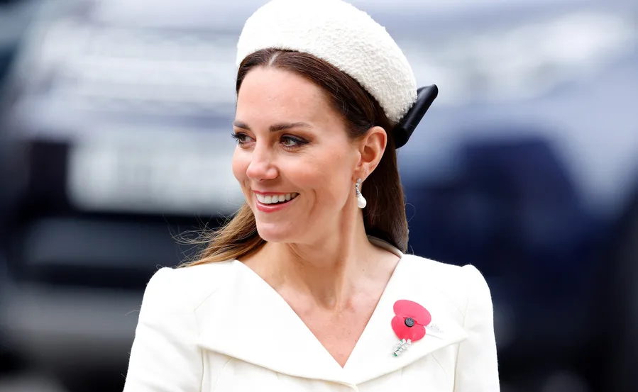 Woman in white dress with poppy pin, smiling, wearing white textured hat and drop earrings.
