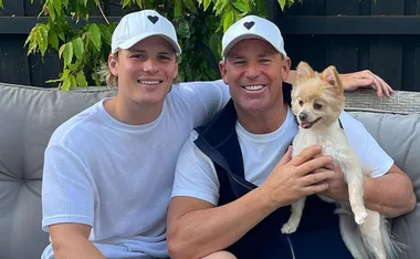 Father and son in white caps sitting on a couch, smiling, holding a small dog outdoors.