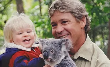 A man and a child smile while holding a koala in an outdoor setting.