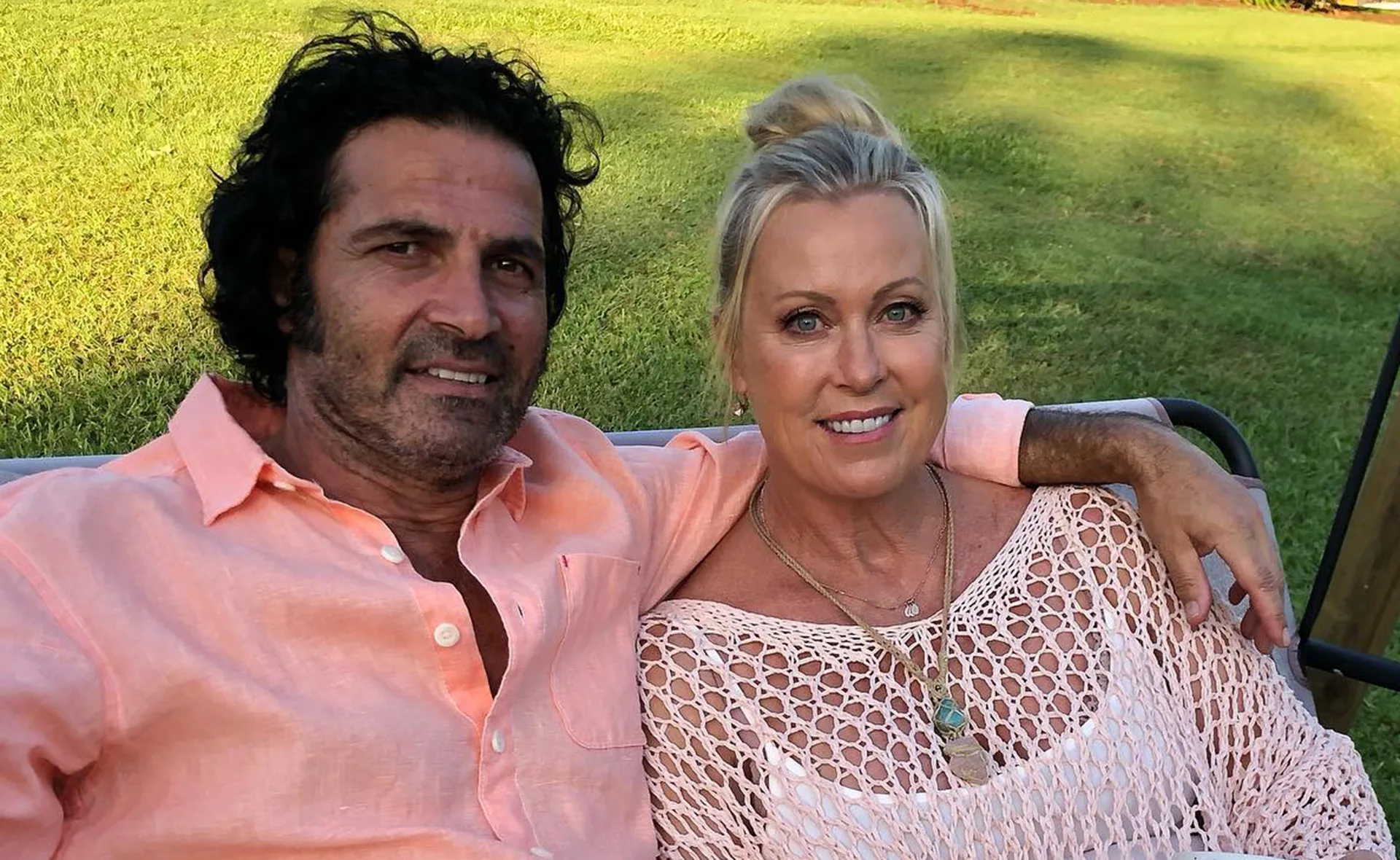 A couple sitting on a bench outdoors, smiling at the camera, with green grass in the background.