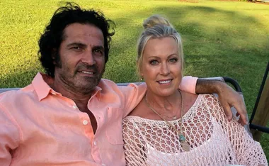 A couple sitting on a bench outdoors, smiling at the camera, with green grass in the background.