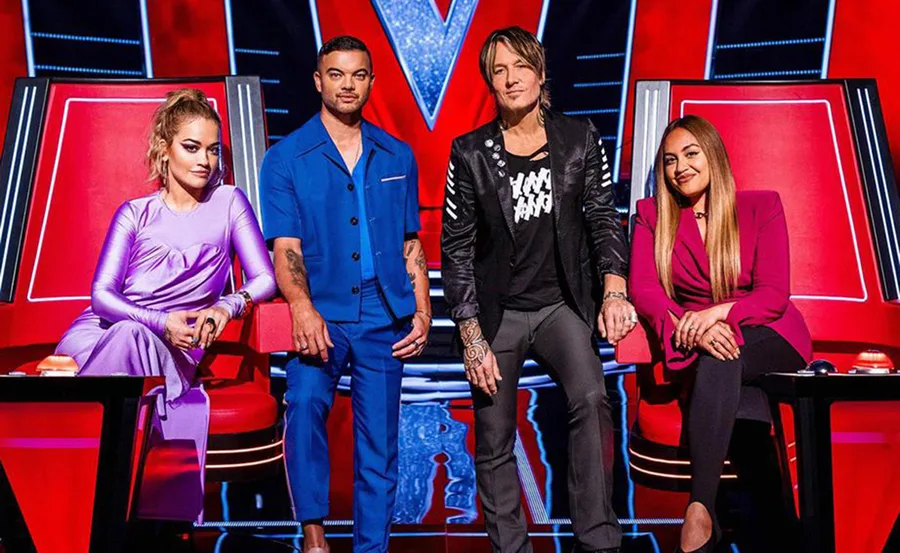 Four judges seated on red chairs on "The Voice," with a large "V" logo in the background.