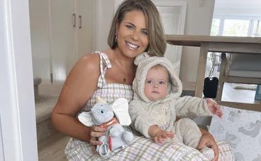 Woman smiling, holding a baby in a fluffy bear onesie, sitting indoors. Baby has a plush elephant toy.