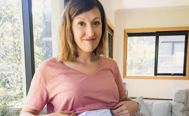 Woman in pink shirt smiling indoors, holding her baby bump near a window.