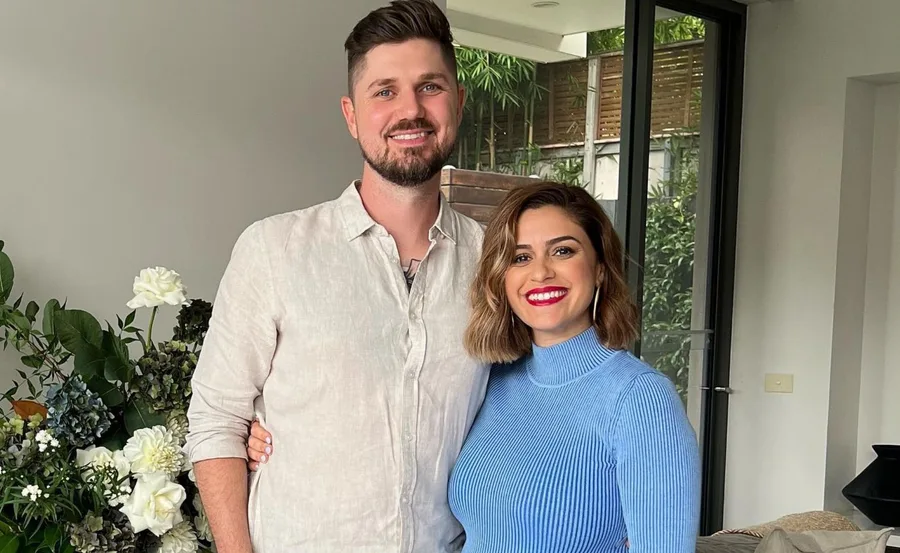 A smiling couple stands indoors in front of a window and greenery, with flowers nearby.