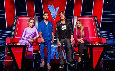 Judges of "The Voice" seated on red chairs with a large "V" logo backdrop.