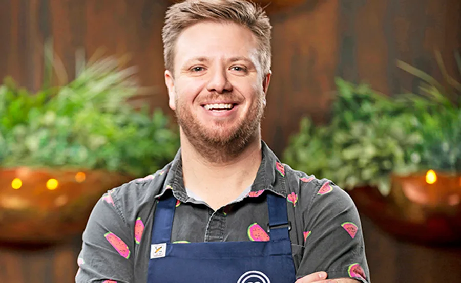 Man in a watermelon-patterned shirt and apron smiling, with green plants blurred in the background.