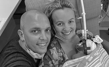 Man and woman smiling, holding a newborn baby wrapped in a blanket with medical equipment attached.