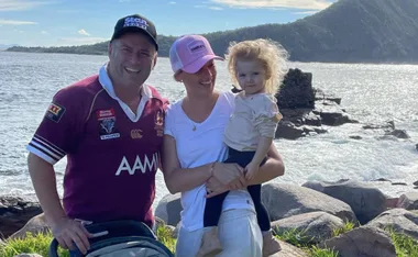 Family smiling near ocean, with a man in a maroon jersey, woman in a pink cap holding a child, rocky shore in background.
