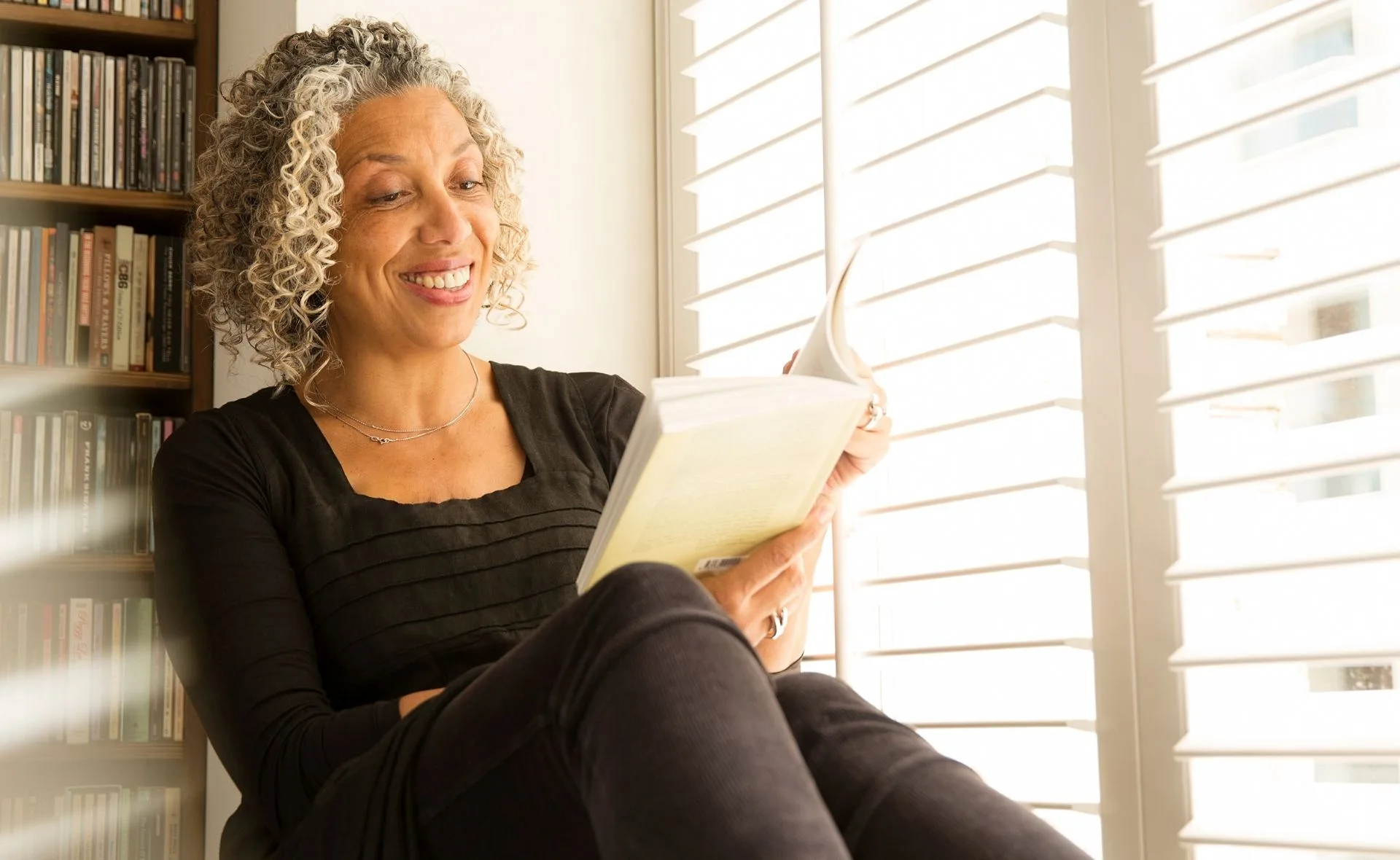 Woman with curly hair in black shirt, smiling while reading a book by a bright window with blinds and a shelf of books.