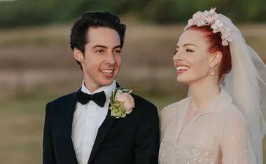 A bride and groom smile at each other outdoors, wearing a wedding dress and suit with a flower boutonniere.