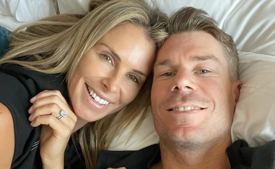 A smiling couple takes a close-up selfie while lying on a bed, showing a ring on one person's hand.