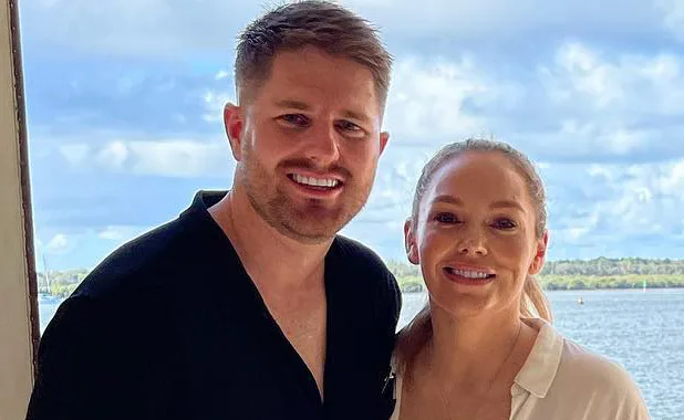 Two people smiling at the camera, standing outdoors with a scenic water and sky background.
