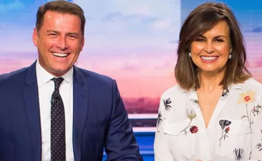 Two TV show hosts smiling on set, one in a blue suit and the other in a floral blouse, with a colorful background.