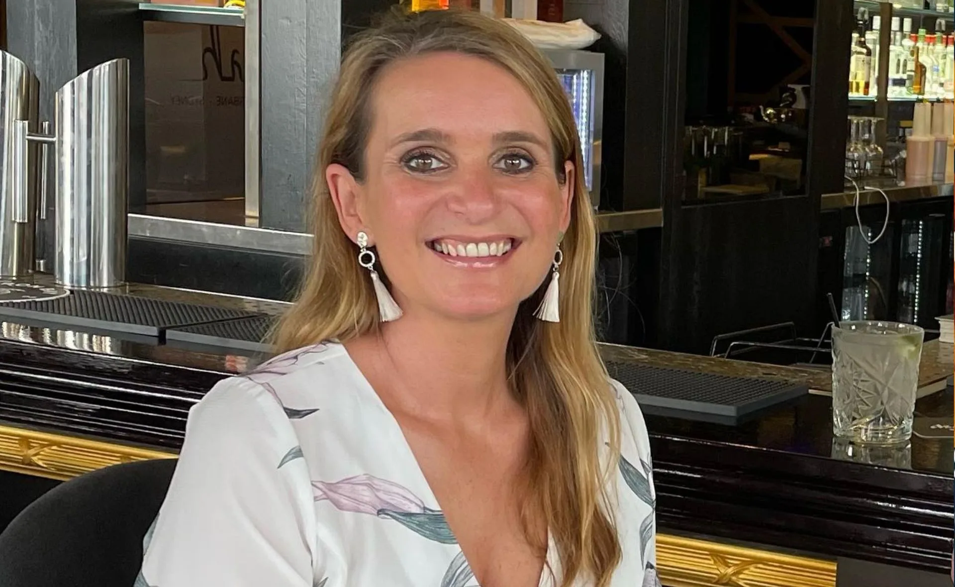 A woman with blond hair and white earrings smiling, sitting at a bar counter with a drink nearby.