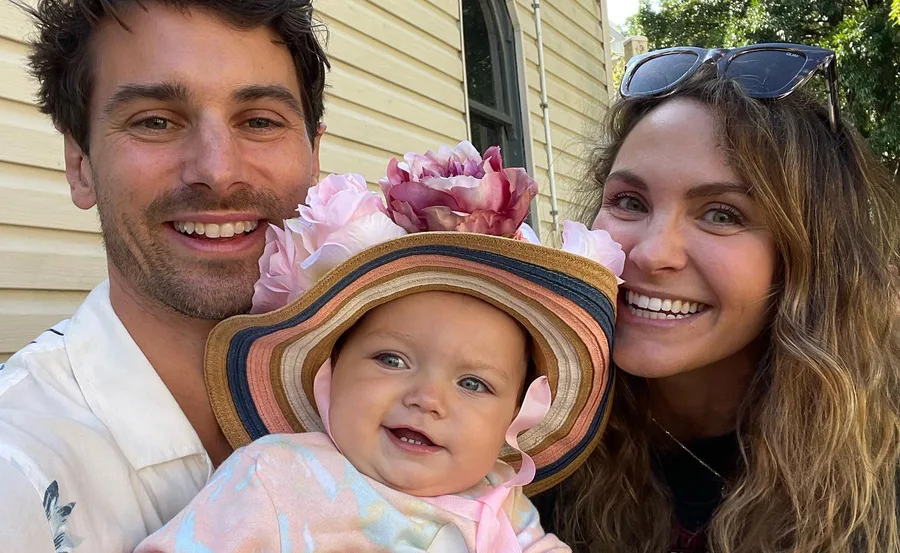 Family smiling with baby in colorful hat and floral headpiece, standing outside near a house.