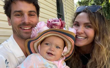 Family smiling with baby in colorful hat and floral headpiece, standing outside near a house.