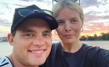 A couple smiling at the camera with a sunset and body of water in the background.
