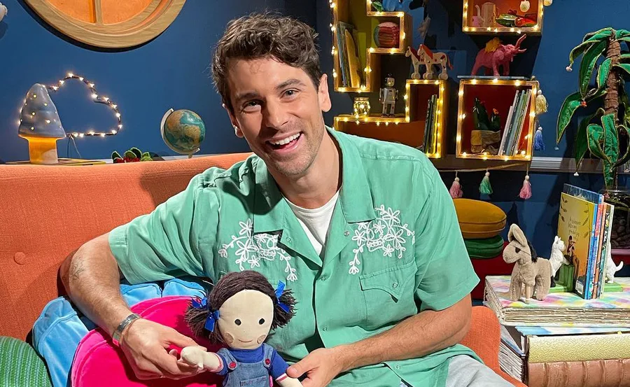 Smiling person in green shirt holding a doll, sitting in a bright, playful room with books and decorations.