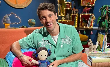 Smiling person in green shirt holding a doll, sitting in a bright, playful room with books and decorations.