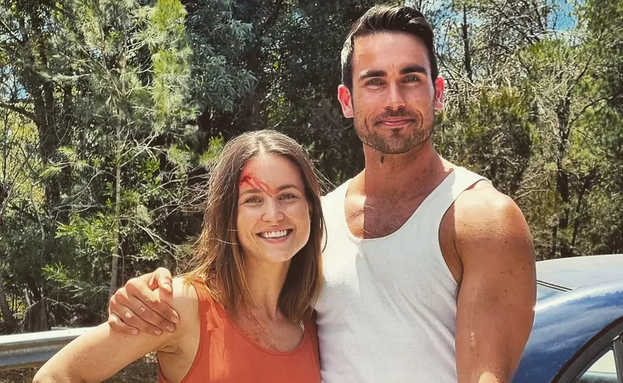 Two people smiling outdoors with trees in the background; one has a red mark on their forehead.