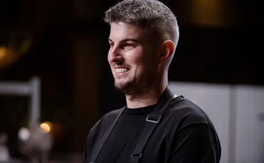 A person in a black apron smiling, standing in a dimly lit room.