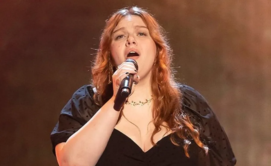 Singer on stage with wavy hair, holding a microphone, wearing a black dress, performing passionately.