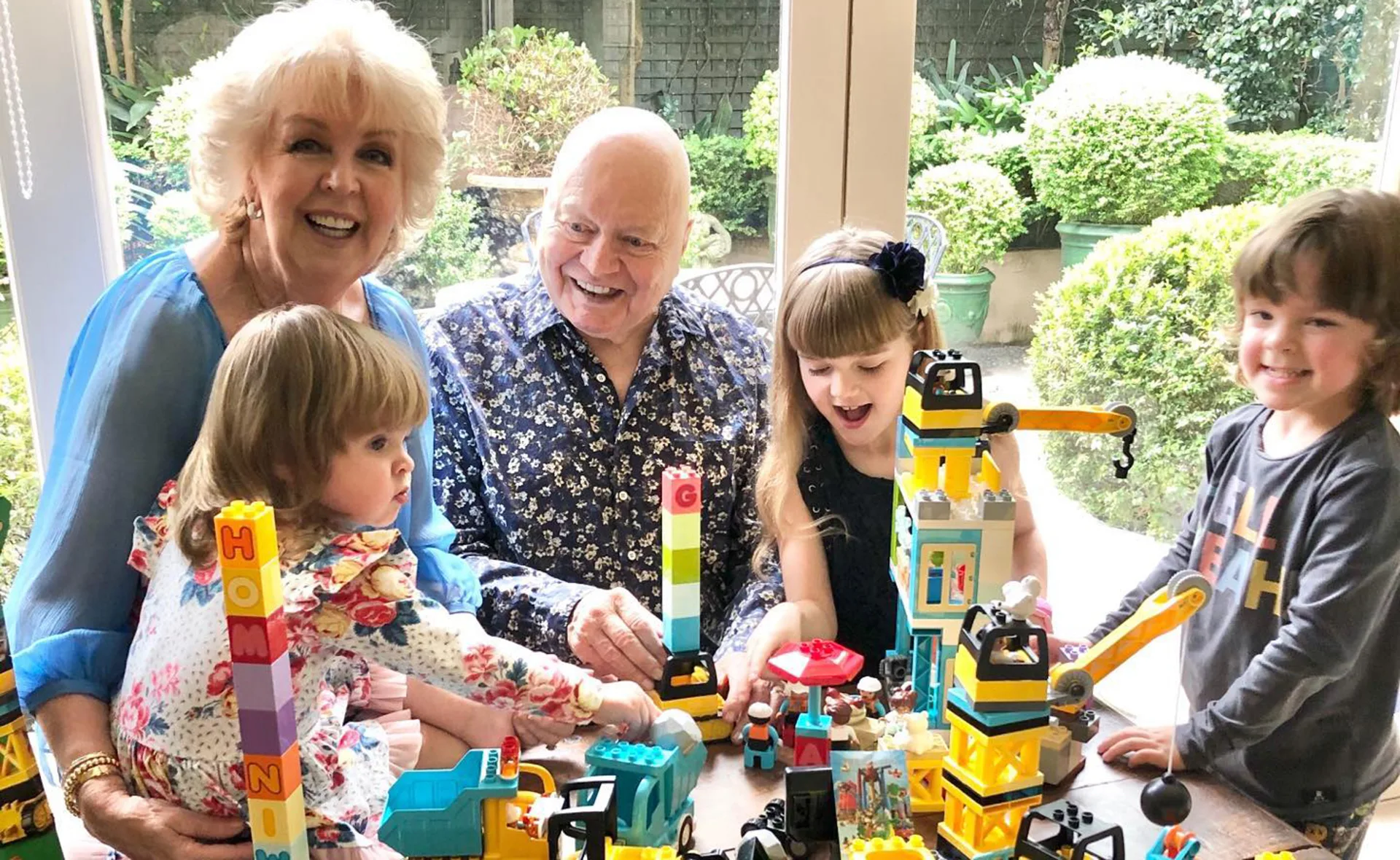 Elderly couple smiling with three young children playing with colorful building blocks at a table.