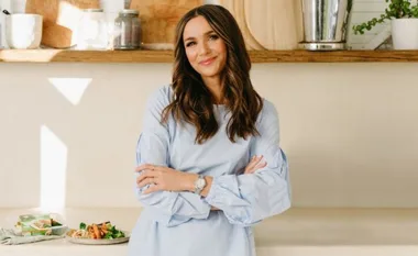 Natalie Gruzlewski standing in a kitchen, wearing a light blue blouse, smiling with arms crossed.