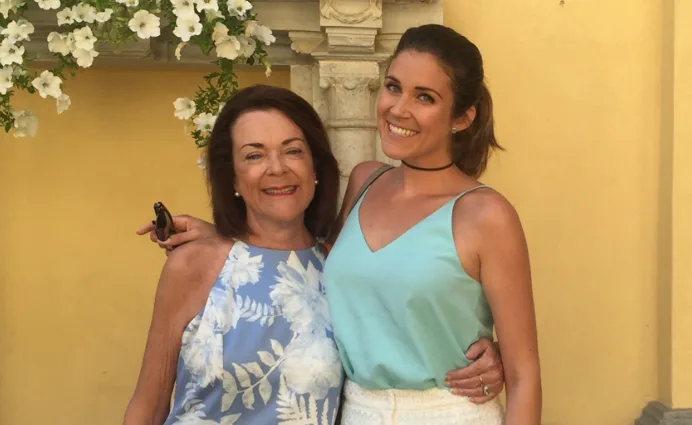Two women smiling and embracing, standing outdoors against a yellow wall adorned with flowers.