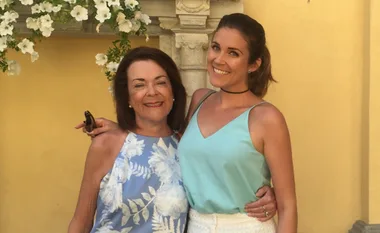 Two women smiling and embracing, standing outdoors against a yellow wall adorned with flowers.