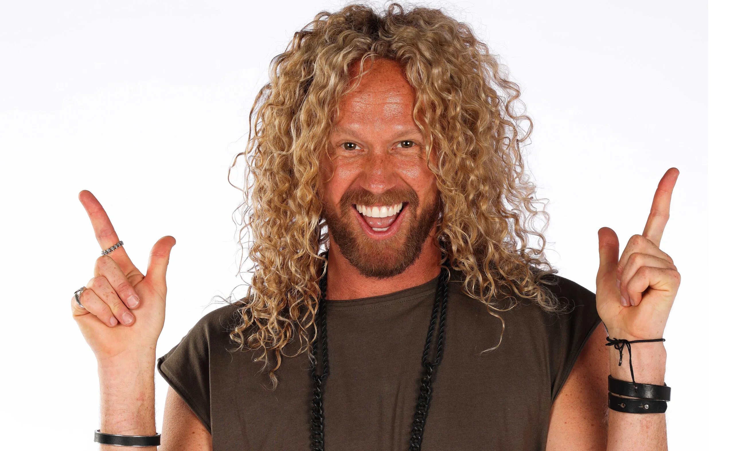 Man with curly blonde hair, smiling broadly, pointing upwards with both hands, wearing casual clothing and leather bracelets.