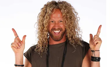 Man with curly blonde hair, smiling broadly, pointing upwards with both hands, wearing casual clothing and leather bracelets.