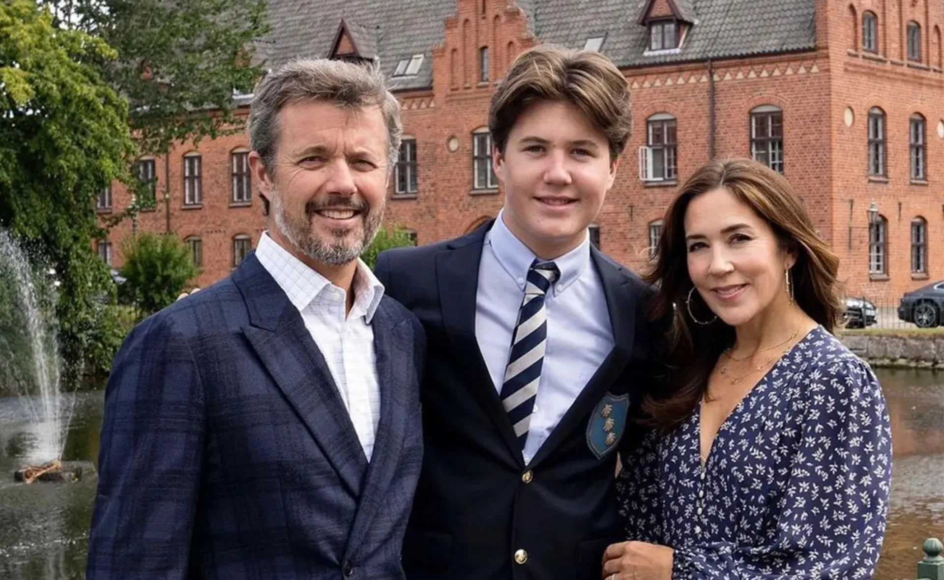 A family stands together smiling, with a brick building in the background.