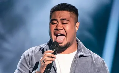 A man passionately singing on stage, holding a microphone, with a spotlighted blue background.