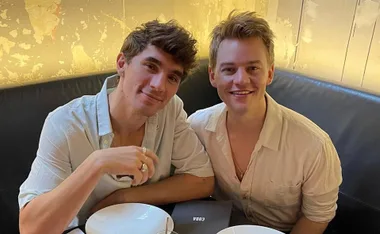 Two people smiling, seated together at a table in a warmly lit setting, wearing casual white shirts.