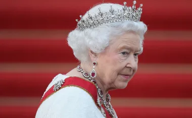 Elderly woman with white hair, wearing a crown and jewelry, against a red background.