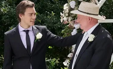 Two men in suits stand together at a floral-decorated outdoor event, one wearing a hat.