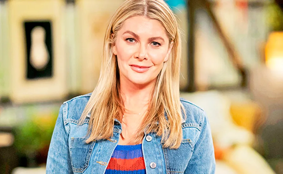 Blonde woman in denim jacket smiling indoors with blurred colorful background.