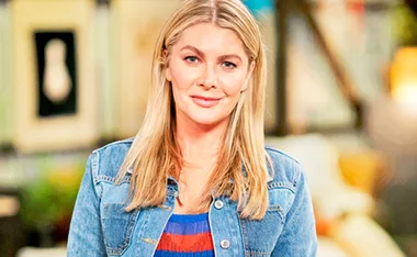 Blonde woman in denim jacket smiling indoors with blurred colorful background.